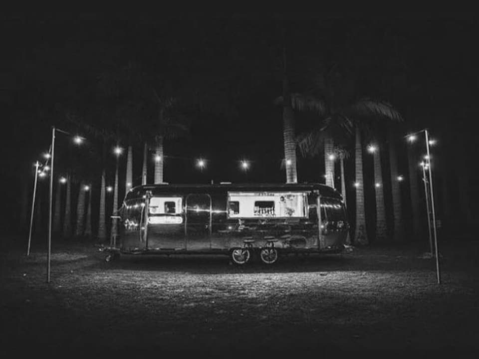 The Airstream glowing at night, framed by palm trees and festoon lighting