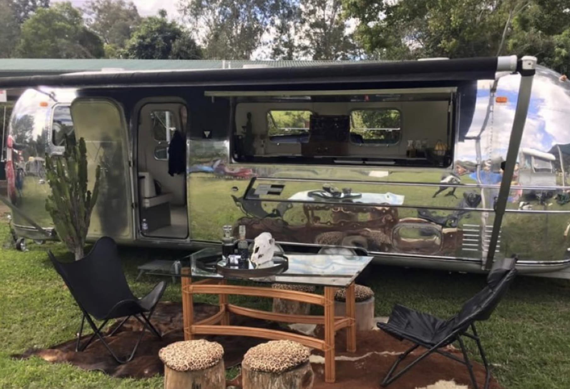 The polished Airstream centred in frame with door open and awning extended, styled with cactus, butterfly chairs and a glass and chrome side table